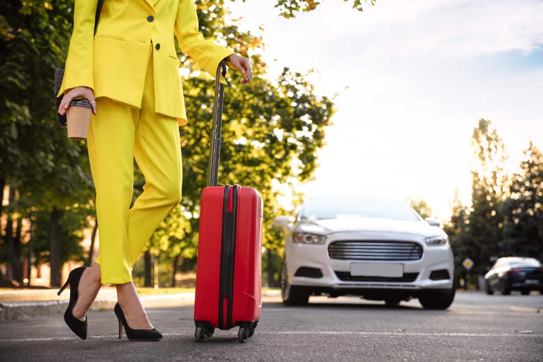 Photo of woman in bright yellow suit with red suitcase waiting for airport car service on city street, closeup