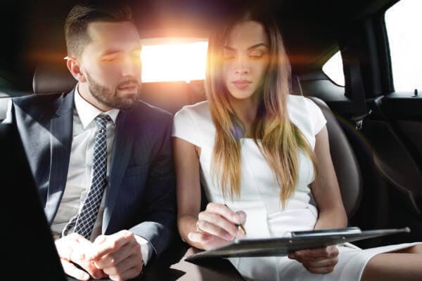 Photo of man and woman working on a project together. They are sitting in the back of a corporate car and working on a tablet and paperwork.