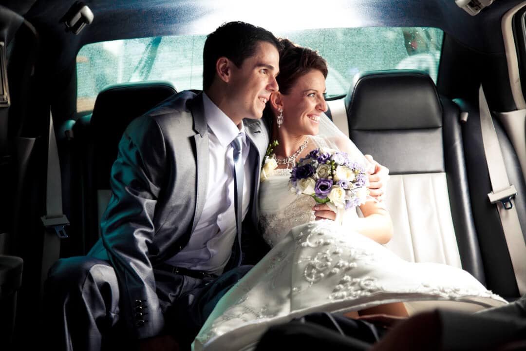 Photo of wedding couple sitting in the back of a luxury car. They are looking at the door that is open and smiling.