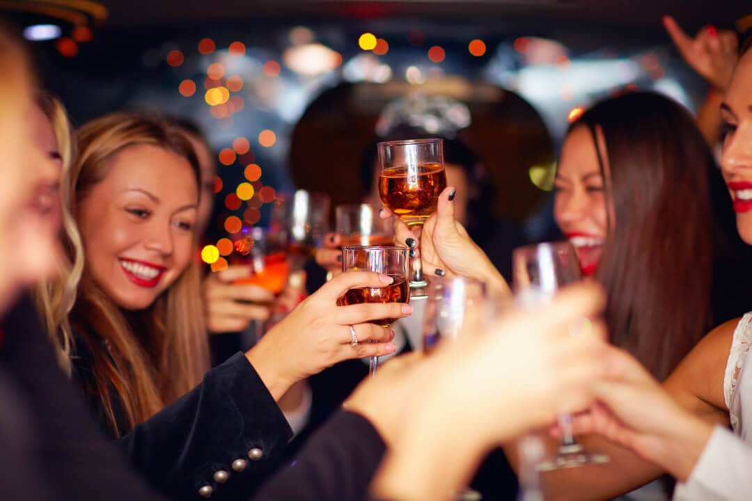 Photo of woman at a bar toasting with wine glasses. They are all smiling and happy. There is twinkle lights in the background.
